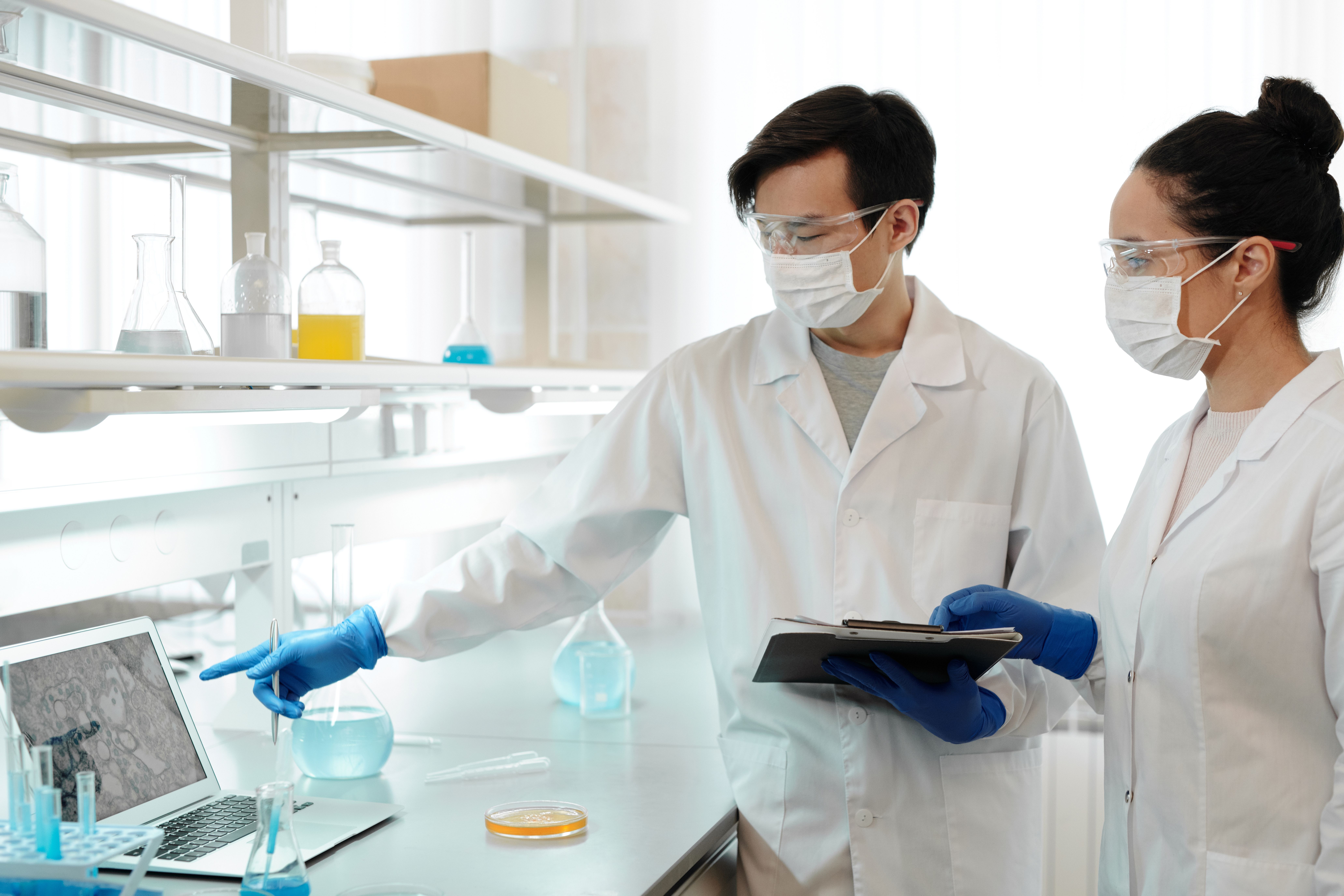 Two scientists in a laboratory reviewing results on a laptop and clipboard